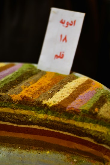 A display of various colorful spices arranged in horizontal layers. Each layer consists of a different spice, creating a vibrant and textured pattern. A sign with text in another language is placed upright in the center.