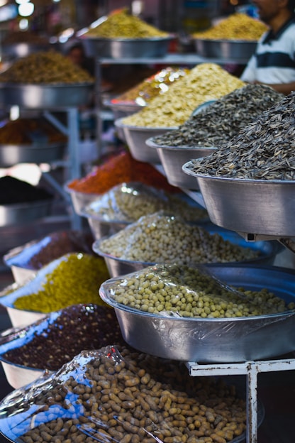 A variety of dried goods and seeds displayed in large metal bowls, each containing different types of grains and legumes. The bowls are organized on multiple shelves, and the scene takes place in what looks like a market or bazaar setting.