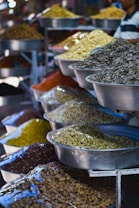 A variety of dried goods and seeds displayed in large metal bowls, each containing different types of grains and legumes. The bowls are organized on multiple shelves, and the scene takes place in what looks like a market or bazaar setting.