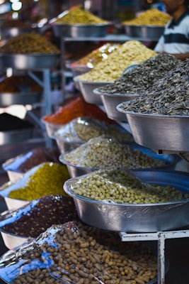 A variety of dried goods and seeds displayed in large metal bowls, each containing different types of grains and legumes. The bowls are organized on multiple shelves, and the scene takes place in what looks like a market or bazaar setting.