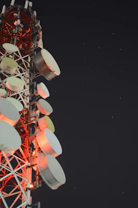 A tall communication tower is equipped with numerous large, round satellite dishes and antennae. The structure is illuminated by red lights against a dark night sky scattered with stars.
