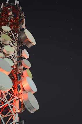 A tall communication tower is equipped with numerous large, round satellite dishes and antennae. The structure is illuminated by red lights against a dark night sky scattered with stars.