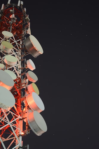 A tall communication tower is equipped with numerous large, round satellite dishes and antennae. The structure is illuminated by red lights against a dark night sky scattered with stars.
