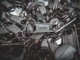 A pile of metallic zipper sliders with visible engraved text on a few of them. The image captures the close-up details and texture of the metal components, highlighting their shiny surfaces.