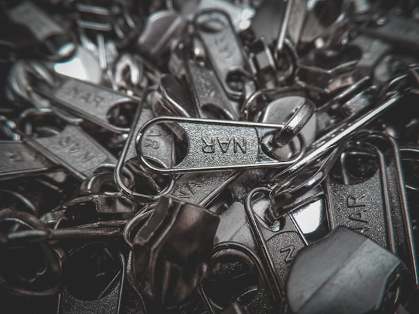 A pile of metallic zipper sliders with visible engraved text on a few of them. The image captures the close-up details and texture of the metal components, highlighting their shiny surfaces.