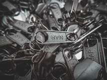 A pile of metallic zipper sliders with visible engraved text on a few of them. The image captures the close-up details and texture of the metal components, highlighting their shiny surfaces.