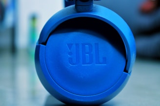 A round blue JBL speaker with a textured surface. The focus is on the JBL logo at the center, and the background is blurred, giving emphasis to the speaker.