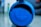 A round blue JBL speaker with a textured surface. The focus is on the JBL logo at the center, and the background is blurred, giving emphasis to the speaker.