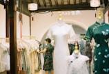 A clothing store featuring traditional dresses displayed on mannequins. A person wearing a traditional dress is selecting an item from a rack filled with various clothing styles. The interior features warm lighting, decorative elements like lanterns, and an arched doorway.