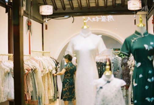 A clothing store featuring traditional dresses displayed on mannequins. A person wearing a traditional dress is selecting an item from a rack filled with various clothing styles. The interior features warm lighting, decorative elements like lanterns, and an arched doorway.