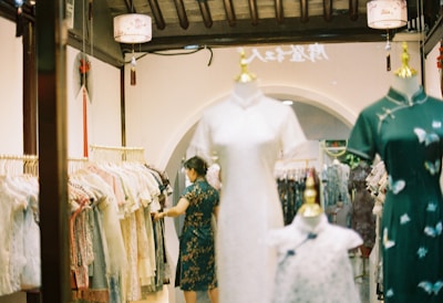 A clothing store featuring traditional dresses displayed on mannequins. A person wearing a traditional dress is selecting an item from a rack filled with various clothing styles. The interior features warm lighting, decorative elements like lanterns, and an arched doorway.