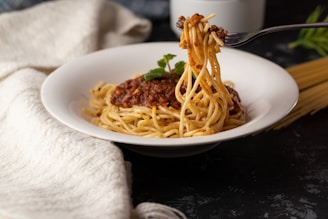 A plate of pasta topped with a rich meat sauce is presented, with a fork twirling the spaghetti. The dish is garnished with a small sprig of fresh mint. A white cloth napkin is draped elegantly to the side, and uncooked spaghetti is visible in the background.