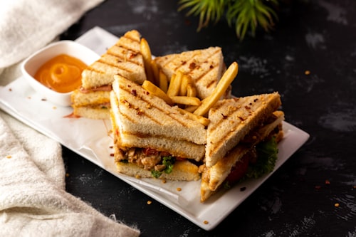 A toasted club sandwich with fries and pot of dipping sauce on a white plate