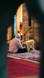 A person is seated on a decorative carpet within a mosque or prayer space, reading from a book placed on a small stand. The setting features ornate pillars and warm lighting.