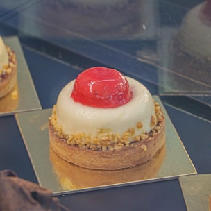 A delicately crafted dessert with a round base of pastry topped with creamy white mousse and adorned with a glossy red jelly sphere. The pastry edges are sprinkled with small nut pieces, and the dessert is presented on a golden square platter.