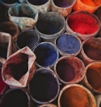Several pots filled with vibrant powder pigments are arranged closely together, showcasing a variety of colors such as red, blue, purple, and orange. The pots appear to be made of clay or similar material, giving a rustic feel to the scene.