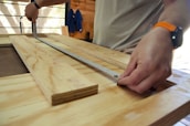 Hands measuring plywood with a metal ruler or measuring tape in a woodworking environment. A person wears a smartwatch and a bright wristband, working on a wooden table.