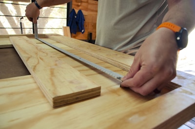 Hands measuring plywood with a metal ruler or measuring tape in a woodworking environment. A person wears a smartwatch and a bright wristband, working on a wooden table.