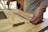 Hands measuring plywood with a metal ruler or measuring tape in a woodworking environment. A person wears a smartwatch and a bright wristband, working on a wooden table.