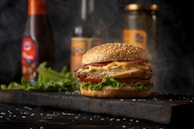 A close-up of a cheeseburger with a sesame seed bun, lettuce, crispy patty, melted cheese, and a slice of tomato. Smoke swirls in the background, suggesting freshly cooked food. Condiment bottles and pickle jars are blurred in the background, placed on a dark wooden surface with scattered sesame seeds.