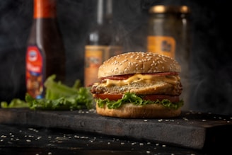 A close-up of a cheeseburger with a sesame seed bun, lettuce, crispy patty, melted cheese, and a slice of tomato. Smoke swirls in the background, suggesting freshly cooked food. Condiment bottles and pickle jars are blurred in the background, placed on a dark wooden surface with scattered sesame seeds.