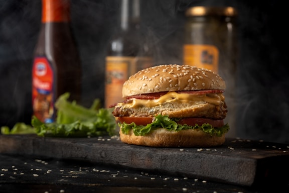 A close-up of a cheeseburger with a sesame seed bun, lettuce, crispy patty, melted cheese, and a slice of tomato. Smoke swirls in the background, suggesting freshly cooked food. Condiment bottles and pickle jars are blurred in the background, placed on a dark wooden surface with scattered sesame seeds.