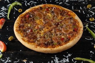 A whole pizza topped with sausage, beef, green olives, and red and green bell peppers on a dark wooden surface. Shredded cheese, sliced green chili peppers, olives, and tomato wedges are scattered around.