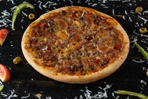 A whole pizza topped with sausage, beef, green olives, and red and green bell peppers on a dark wooden surface. Shredded cheese, sliced green chili peppers, olives, and tomato wedges are scattered around.