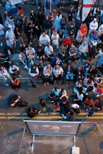 A large group of people gathered outdoors, watching a screen that is displaying a man. The crowd is diverse in age and appears interested and engaged, with many sitting closely together, some standing. Many are dressed casually, some in sports attire, and a few waving or holding flags. The event seems to have a communal, festive atmosphere.