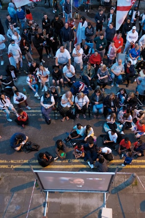 A large group of people gathered outdoors, watching a screen that is displaying a man. The crowd is diverse in age and appears interested and engaged, with many sitting closely together, some standing. Many are dressed casually, some in sports attire, and a few waving or holding flags. The event seems to have a communal, festive atmosphere.