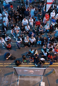 A large group of people gathered outdoors, watching a screen that is displaying a man. The crowd is diverse in age and appears interested and engaged, with many sitting closely together, some standing. Many are dressed casually, some in sports attire, and a few waving or holding flags. The event seems to have a communal, festive atmosphere.