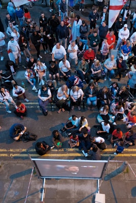 A large group of people gathered outdoors, watching a screen that is displaying a man. The crowd is diverse in age and appears interested and engaged, with many sitting closely together, some standing. Many are dressed casually, some in sports attire, and a few waving or holding flags. The event seems to have a communal, festive atmosphere.