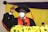 An individual is wearing academic regalia, including a black gown, red stole, and a graduation cap with a tassel. They are speaking into a microphone while making a fist gesture. The background features draped yellow and purple fabric, and there is a decorative arrangement of yellow flowers in the foreground.