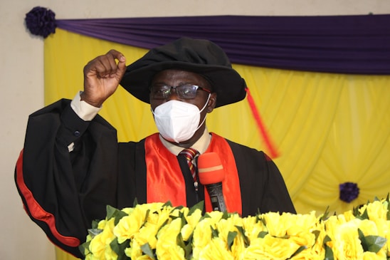 An individual is wearing academic regalia, including a black gown, red stole, and a graduation cap with a tassel. They are speaking into a microphone while making a fist gesture. The background features draped yellow and purple fabric, and there is a decorative arrangement of yellow flowers in the foreground.