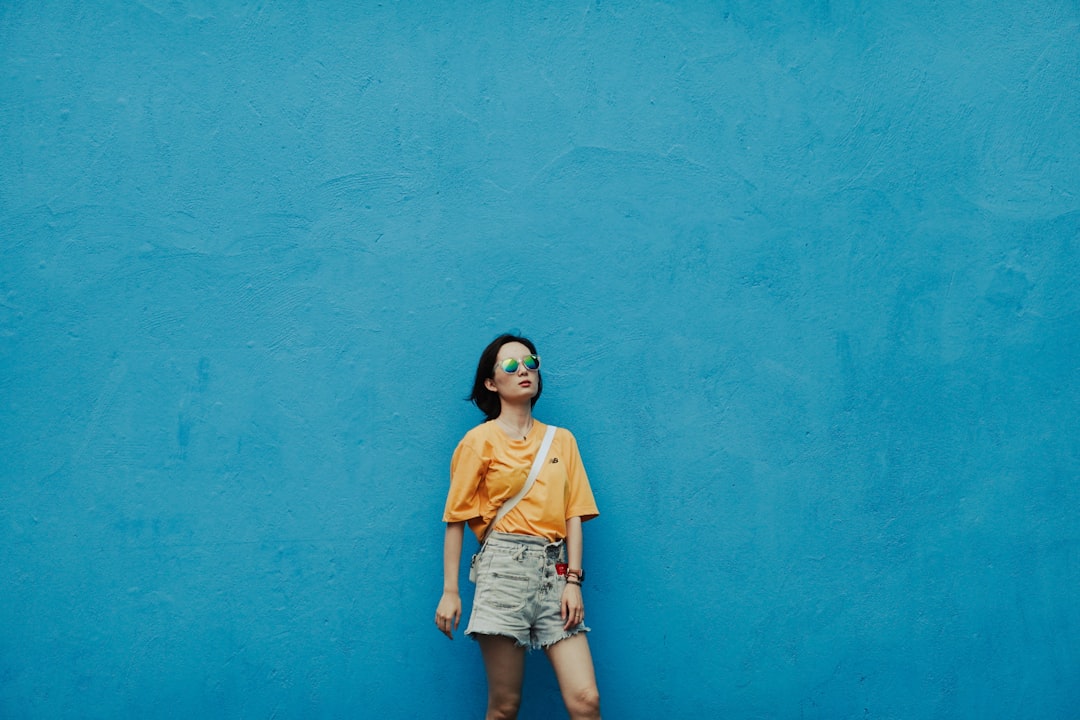 woman in yellow t-shirt and blue denim shorts standing beside blue wall