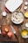 A kitchen scene with ingredients and tools arranged on a wooden table. A cutting board holds three apples and a knife. A bowl contains oatmeal mixed with dried cranberries, accompanied by a metal spatula. Nearby, a small saucepan holds a yellow liquid, possibly melted butter, with a whisk in it. An egg carton sits opened with one cracked shell visible, and a baking dish with a red handle is ready for use.