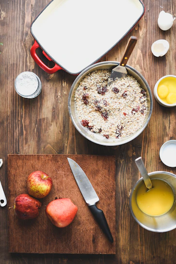 A kitchen scene with ingredients and tools arranged on a wooden table. A cutting board holds three apples and a knife. A bowl contains oatmeal mixed with dried cranberries, accompanied by a metal spatula. Nearby, a small saucepan holds a yellow liquid, possibly melted butter, with a whisk in it. An egg carton sits opened with one cracked shell visible, and a baking dish with a red handle is ready for use.