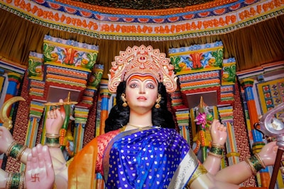 A colorful and detailed statue of a multi-armed deity in a traditional Indian temple setting, adorned with intricate jewelry and a decorated headdress. The statue is set against a richly patterned and ornately carved backdrop with vibrant colors and motifs.