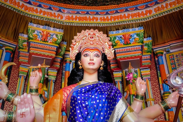 A colorful and detailed statue of a multi-armed deity in a traditional Indian temple setting, adorned with intricate jewelry and a decorated headdress. The statue is set against a richly patterned and ornately carved backdrop with vibrant colors and motifs.