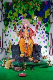 A statue adorned in vibrant traditional attire sits on a black rock pedestal. The figure is embellished with a detailed headdress and holds a trident and a pot. Surrounding the statue are various offerings, including plates, bowls, and decorative items. The backdrop features colorful leaves in shades of green, blue, and yellow, creating a festive atmosphere.