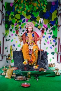 A statue adorned in vibrant traditional attire sits on a black rock pedestal. The figure is embellished with a detailed headdress and holds a trident and a pot. Surrounding the statue are various offerings, including plates, bowls, and decorative items. The backdrop features colorful leaves in shades of green, blue, and yellow, creating a festive atmosphere.