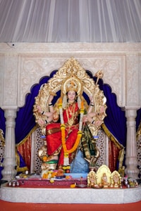 A detailed and ornate statue of a Hindu goddess is seated on a decorative throne surrounded by intricate carvings. The goddess wears vibrant red and green garments, adorned with garlands of orange and white flowers and jewelry. She has multiple arms, holding various symbolic items, and a serene expression on her face. The backdrop consists of deep blue drapes contrasted with intricate white paneling. On the platform below are offerings and smaller golden figures.