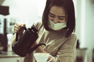 A person wearing a mask is carefully pouring liquid from a black kettle into a paper filter, likely in a coffee-making process. The setting appears to be indoors with a blurred background, suggesting a kitchen or café.