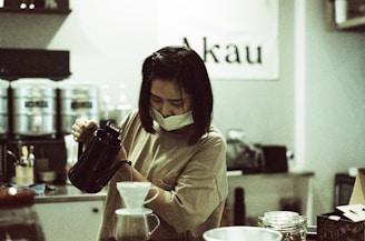 A person wearing a face mask is focused on pouring hot water from a kettle into a pour-over coffee maker. The setting is a cozy cafe with a sign reading 'Akau' in the background. Various coffee equipment and jars are visible on the counter.