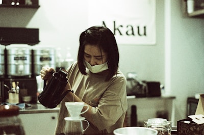A person wearing a face mask is focused on pouring hot water from a kettle into a pour-over coffee maker. The setting is a cozy cafe with a sign reading 'Akau' in the background. Various coffee equipment and jars are visible on the counter.