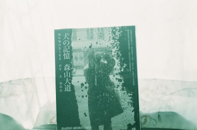 A book cover features the title 'MEMORIES OF A DOG' along with Japanese text. The central image depicts a silhouette of a person in a coat taking a photograph, surrounded by an abstract pattern reminiscent of splattered ink or paint. The overall color scheme is green, contributing to a vintage or artistic look.