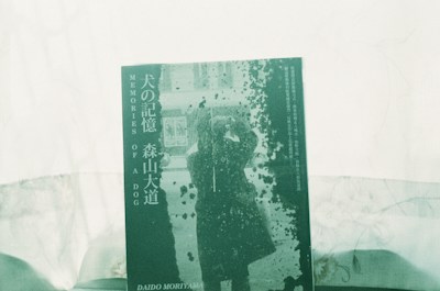 A book cover features the title 'MEMORIES OF A DOG' along with Japanese text. The central image depicts a silhouette of a person in a coat taking a photograph, surrounded by an abstract pattern reminiscent of splattered ink or paint. The overall color scheme is green, contributing to a vintage or artistic look.