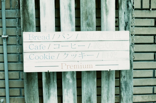 A wooden sign is affixed to a rustic wooden fence with a background of brickwork. The sign features text in both English and Japanese, listing items such as bread, cafe, and cookie. The word 'Premium' is highlighted at the bottom.