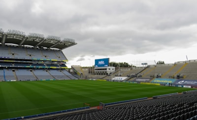A large, empty sports stadium features rows of blue and gray seats surrounding a well-maintained green grass field. The stadium has a modern design with an overhanging roof on one side providing partial cover. Advertising banners are visible along the seating areas, and a large screen displays text. The sky above is overcast, casting a soft light across the scene.