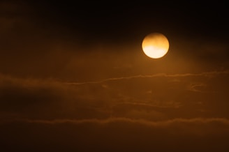 A glowing celestial object, likely the sun or moon, is partially obscured by thin, wispy clouds. The sky is a gradient of dark and light shades, creating a dramatic and serene atmosphere.